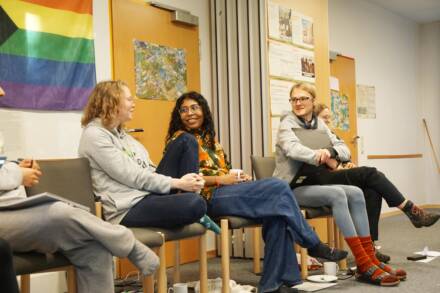 Gruppe diskutiert vor Regenbogenflagge im Raum.