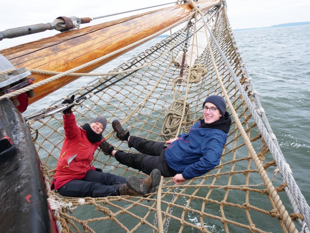 Zwei Personen im Netz eines Segelschiffs lächeln.