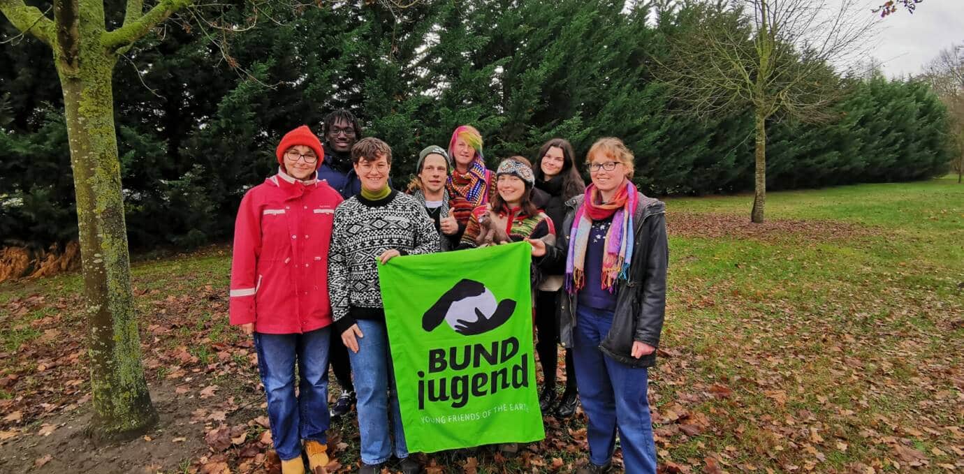 Mehrere Teamer*innen stehen draußen auf einer Streuobstwiese und halten feierlich eine BUNDjugend-Fahne und eine Wildkatze hoch.