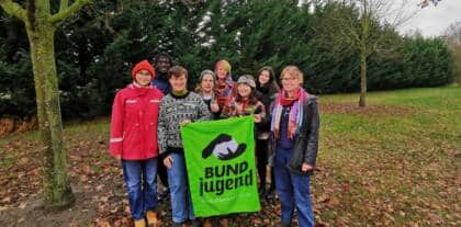 Mehrere Teamer*innen stehen draußen auf einer Streuobstwiese und halten feierlich eine BUNDjugend-Fahne und eine Wildkatze hoch.